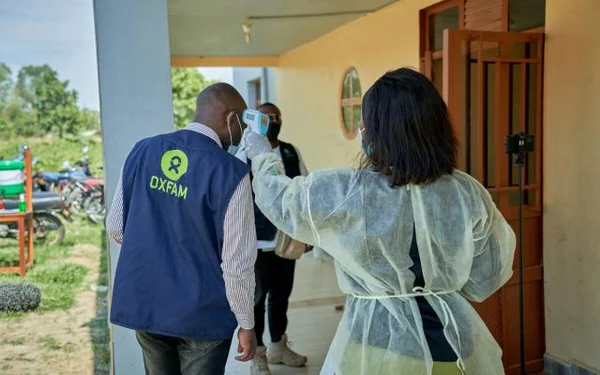 Un agent de santé en blouse de protection prend la température de Roger Kalimira, vêtu d’un gilet bleu Oxfam, à l’entrée d’une réunion communautaire dans la zone de santé de Rwampara, en RDC, lors de la lutte contre Ebola.