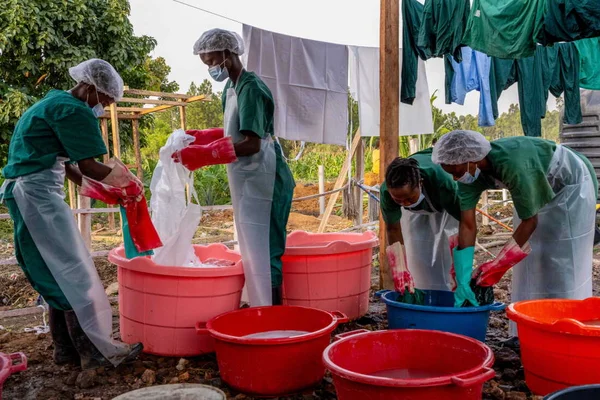 Quatre agents d’hygiène en blouses vertes, tabliers blancs, charlottes et masques lavent des équipements de protection individuelle dans des bassines en plastique à la buanderie du centre de traitement Ebola de Rwampara, en Ituri, dans l’est de la RDC.