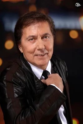Portrait de Frank Michael souriant légèrement, vêtu d'un jean noir sur une chemise blanche et un nœud noir, contre un fond flou avec des lumières bokeh