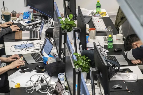 Photographie d'un espace de travail de bureau encombré avec plusieurs écrans d'ordinateur et ordinateurs portables, montrant une personne travaillant tandis qu'une autre dorme la tête sur le bureau.