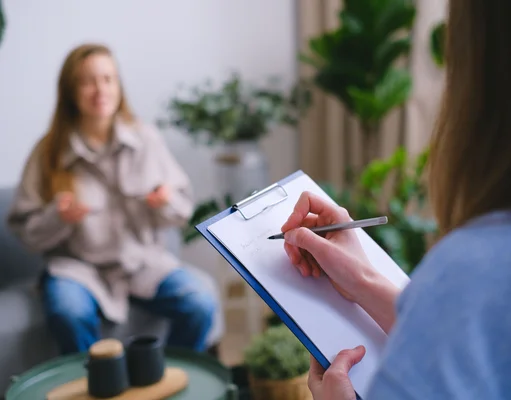 Personne prenant des notes sur un dossier pendant qu'une femme écoute assise sur un canapé dans une pière avec des plantes, dans le cadre de Mon Soutien Psy