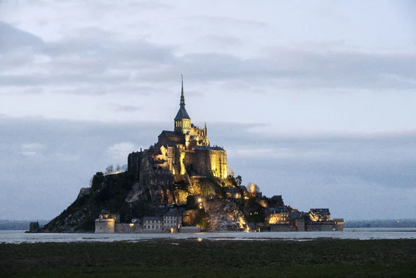 Photographie du Mont-Saint-Michel à l'aube, avec l'abbaye éclairée par des éclats de lumière dorée, dominée par une coupole médiévale et entourée de marais et d'eau