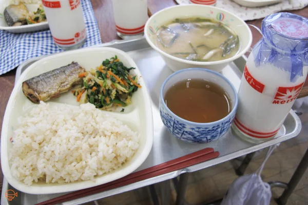 Repas scolaire japonais servi sur un plateau métallique, comprenant du riz blanc, du poisson grillé, des légumes, de la soupe miso, du thé vert et du lait