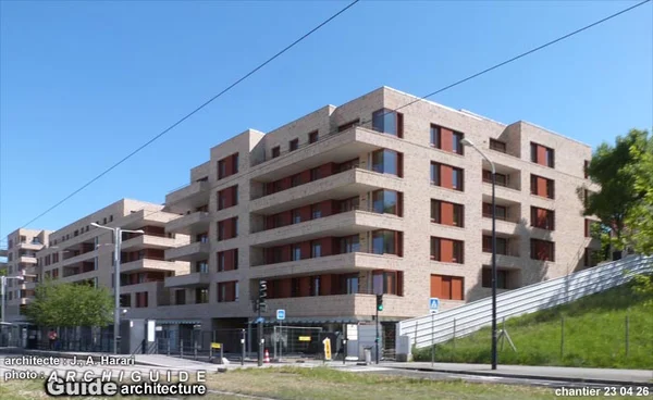 Immeuble résidentiel moderne de plusieurs étages avec facade graduée et balcons, situé sur un coin de rue. Facade en brique claire avec cadres de fenêtres marron-rouge, pente herbeuse et mur de soutènement à droite.