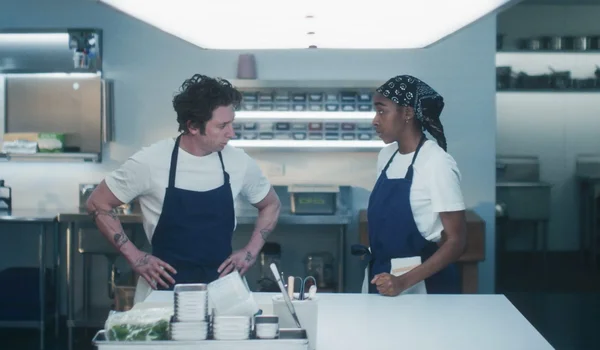Un homme et une femme en chemise blanche et tablier bleu dans une cuisine professionnelle, évoquant l'équipe son de The Bear et l'ambiance sonore stressante du restaurant.