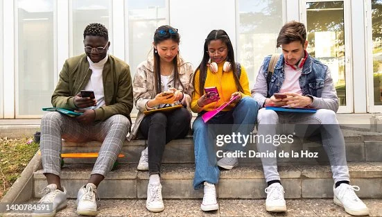 Quatre jeunes étudiants multiculturels assis sur des marches en béton en extérieur, chacun absorbé par son smartphone tout en tenant cahiers ou livres