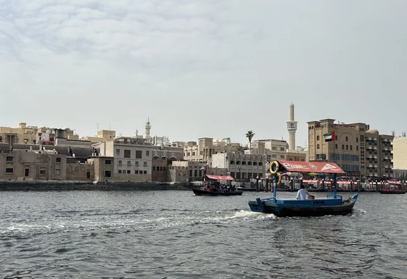 Le Dubai Creek avec un abra traditionnel bleu traversant l'eau au premier plan, face aux quartiers historiques d'Al Shindagha et Al Fahidi avec minarets et tours à vent sous un ciel couvert