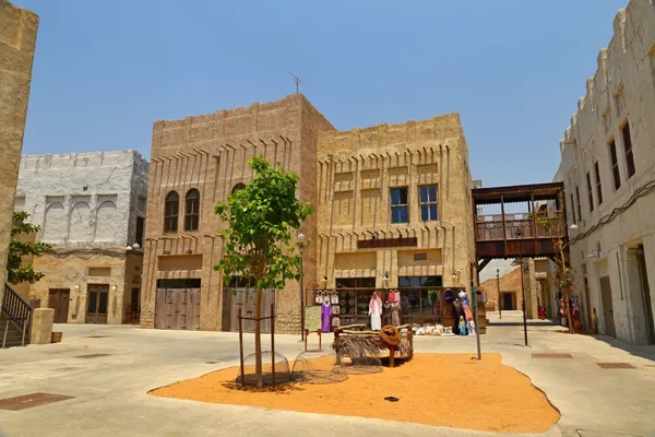 Ruelle du quartier historique Al Fahidi à Dubaï avec bâtiments traditionnels couleur sable aux façades décorées, arbre central, vêtements suspendus en vente et passerelle piétonne en bois au-dessus de la rue
