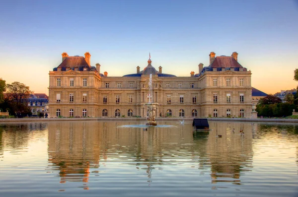 Le palais du Luxembourg, siège du Sénat à Paris, avec sa façade illuminée et la fontaine se reflétant dans l'eau calme du Grand Bassin sous un ciel au coucher du soleil
