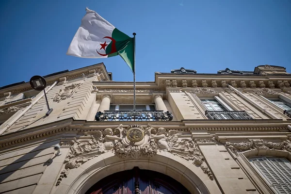 Drapeau algérien vu en contre-plongée flottant au mât au-dessus de la façade classique en pierre ornée de chérubins sculptés, de colonnes et d'une porte cintrée, sous un ciel bleu limpide