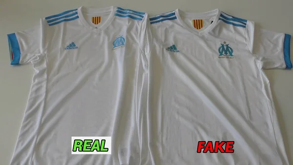 Comparaison côte à côte de deux maillots blancs Adidas de l'Olympique de Marseille, celui de gauche étiqueté REAL et celui de droite FAKE pour montrer les différences entre authentique et contrefaçon