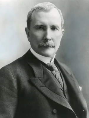 Portrait en noir et blanc de John D. Rockefeller d'âge moyen, moustache épaisse, portant un costume trois-pièces sombre avec cravate à motifs et col rigide, regardant fixement l'objectif
