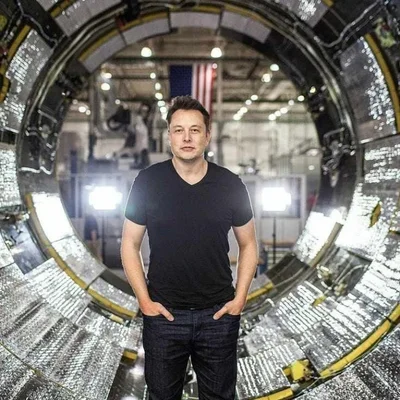 Elon Musk en t-shirt noir à col en V et jean, mains dans les poches, debout dans un grand composant aérospatial circulaire avec un drapeau américain en arrière-plan