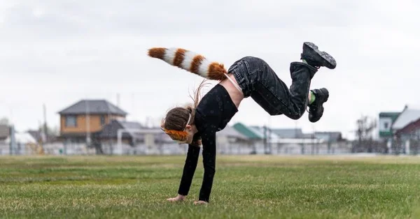 Enfant pratiquant le quadrobics effectuant un salto arrière sur une pelouse, vêtu d'un haut noir et d'un pantalon sombre avec une queue touffue rayée de panda roux attachée à la taille
