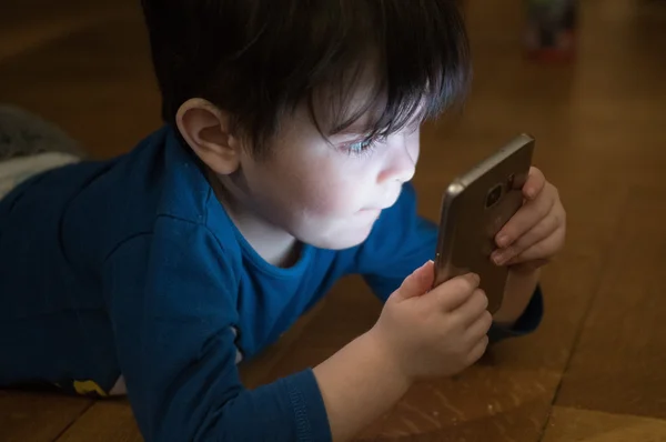 Un jeune enfant allongé sur un parquet en bois, tenant un smartphone dont l'écran illumine son visage qu'il fixe intensément, illustration de l'exposition précoce à TikTok