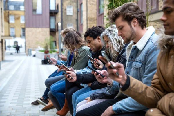 Un groupe d'adolescents assis côte à côte en extérieur sur une allée pavée, chacun penché sur son smartphone pour échanger des messages sur les réseaux sociaux
