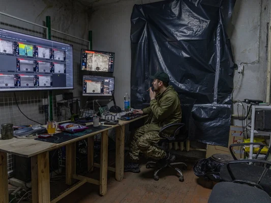 Un homme en veste militaire verte, pantalon camouflage et casquette fumant devant plusieurs écrans de surveillance dans un poste de commandement secret de drones ukrainien aux murs en béton
