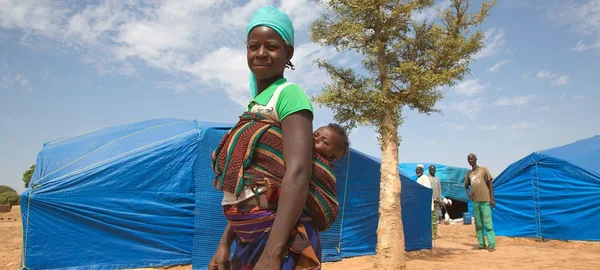 Une femme en chemise verte et foulard bleu portant un bébé sur le dos dans un tissu coloré, debout devant des tentes en bâche bleue dans un camp de déplacés du Sahel central sous un ciel nuageux