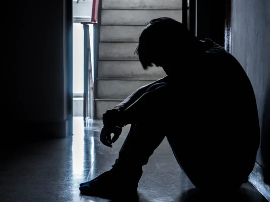 Silhouette d'un adolescent assis recroquevillé contre un mur dans un escalier sombre, tête baissée, illustrant le mal-être et la détresse des jeunes face au monde
