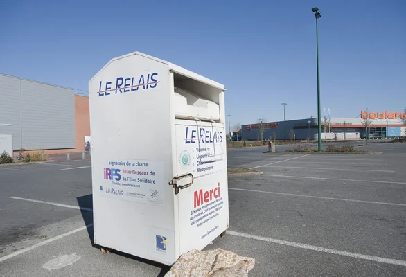 Une benne de collecte de vêtements usagés appartenant à l'organisation Le Relais, stationnée dans un parking vide sous un ciel ensoleillé, entourée de bâtiments commerciaux.
