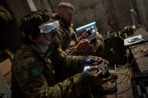 Deux soldats en uniformes camouflés pilotant des drones FPV dans un bunker souterrain sombre en Ukraine, celui au premier plan portant des lunettes FPV et tenant une radiocommande, le second derrière avec ordinateur portable