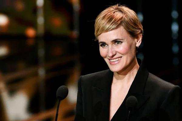 Judith Godrèche, cheveux courts blonds, en blazer noir, souriant derrière deux micros lors de son audition au Sénat sur les violences sexuelles dans le cinéma.