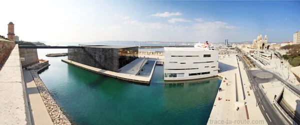 Image panoramique générée par IA d'une ville côtière futuriste avec une architecture moderne, des canaux et un pont sous un ciel lumineux, avec le filigrane trace-ta-route.com.