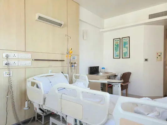 Chambre d'hôpital moderne avec lit médicalisé réglable aux draps blancs, table de chevet, chaise et équipements médicaux fixés au mur.
