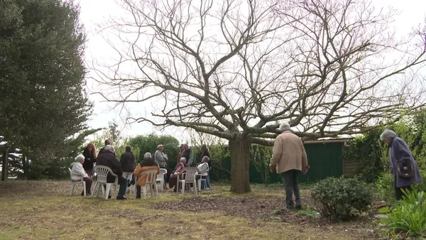 Collectif de riverains réunis en séminaire dans un jardin sous un grand arbre découvert, assis sur des chaises blanches, certains debout à proximité.