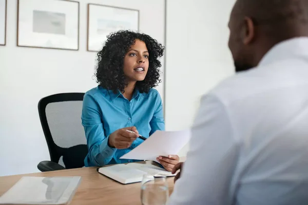Une femme aux cheveux bruns bouclés, vêtue d'une chemise bleue, assise à un bureau avec un stylo et une feuille, échange avec un homme de dos dans un cadre professionnel.