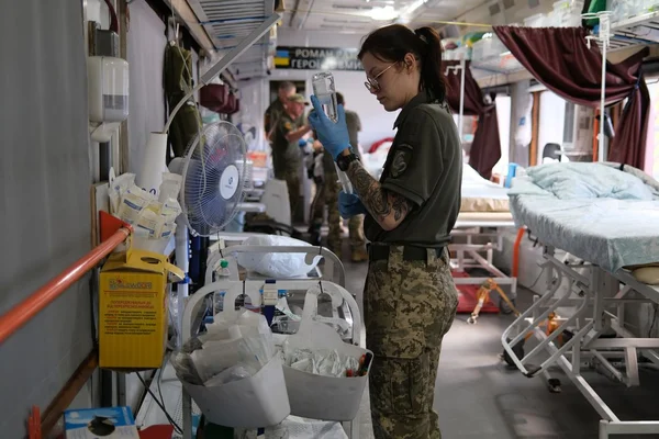 Une soignante en uniforme militaire et gants bleus prépare une perfusion à bord d'un train-hôpital ukrainien évacuant des soldats blessés du front, avec des lits et du personnel en arrière-plan.