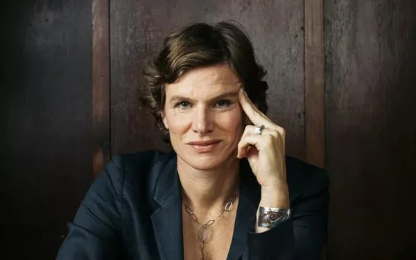 Portrait de Mariana Mazzucato, économiste aux cheveux bruns courts et ondulés, vêtue d'une veste sombre et de bijoux, la main posée contre sa tempe sur un fond sombre texturé.