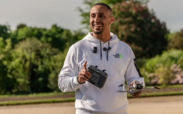 Amine aux cheveux courts et barbe taillée, souriant en sweat blanc à capuche, tenant un drone gris et sa télécommande dans un parc
