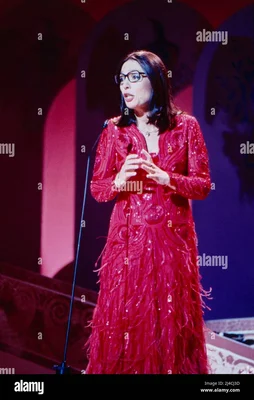 Nana Mouskouri, chanteuse grecque à cheveux sombres et lunettes, en robe rouge scintillante à franges, chantant au micro sur une scène éclairée en bleu et violet lors d'une émission télévisée en Allemagne vers 1980