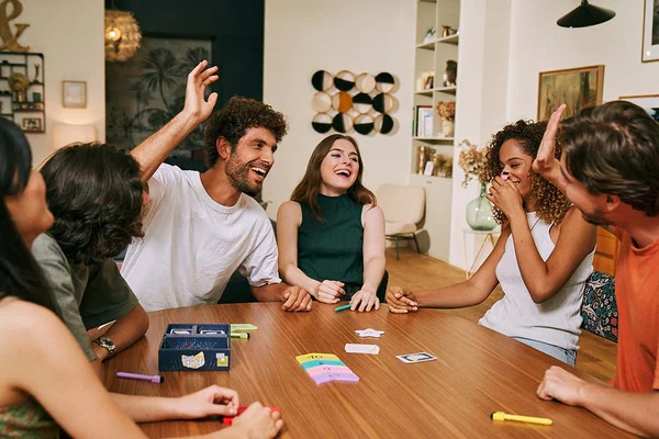 Des jeunes adultes divers rient autour d'une table en bois en jouant à Fun Facts, un jeu de société de Repos Production, dans une pièce moderne et lumineuse.