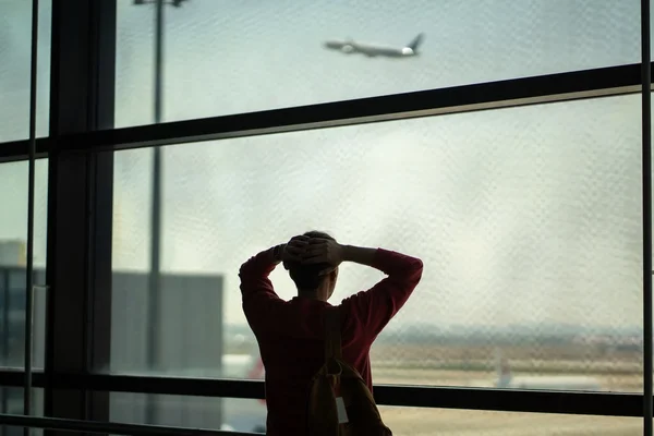 Personne en chemise rouge regardant à travers une fenêtre d'aéroport, avec un avion visible sur la piste