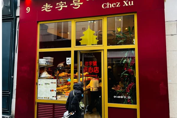 Devanture d'un commerce à la façade rouge et aux encadrements de fenêtres jaunes, portant des caractères chinois et l'enseigne « Chez Xu », avec une personne debout près de l'entrée, dans le quartier de Little Wenzhou à Paris.