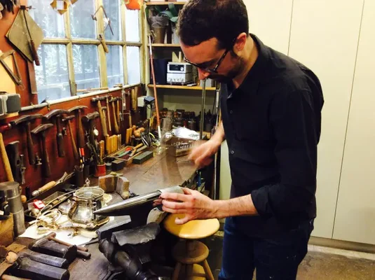 Un artisan orfèvre à lunettes et barbe travaillant dans l'atelier Richard Orfèvres, un atelier figé dans le temps, entouré d'outils et d'équipements.