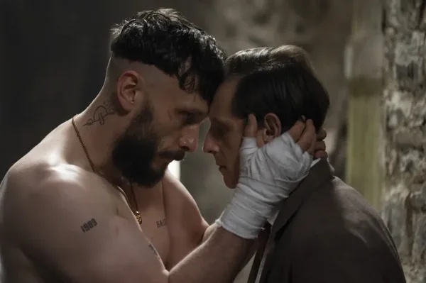 Richard Gadd en Ruben, torse nu, barbu et tatoué à la main bandée, tenant le visage de Jamie Bell en Niall en costume, fronts collés contre un mur en pierre dans Half Man