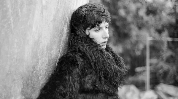 Aldous Harding aux cheveux courts bouclés, portant un manteau sombre texturé, l'épaule appuyée contre un mur en béton clair et le regard tourné sur le côté, en noir et blanc