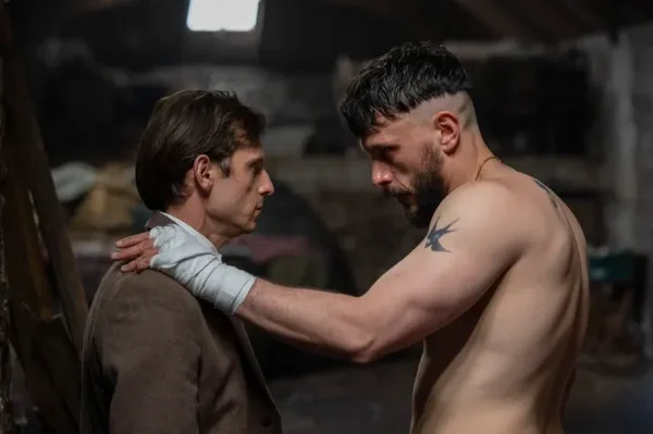 Jamie Bell et Richard Gadd dans Half Man : un homme torse nu, barbu et musclé, main bandée et tatouage d'hirondelle sur l'épaule, posant la main sur un jeune homme en veste marron dans un intérieur rustique sombre