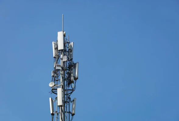 Vue rapprochée de la partie supérieure d'une station de base de télécommunications 5G avec antennes panneaux blanches, boîtiers, câbles et paratonnerre sur mât en acier sous ciel bleu dégagé