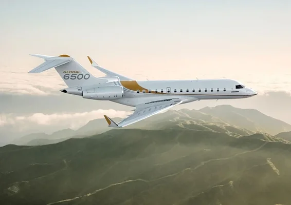 Jet d'affaires Bombardier Global 6500 blanc avec accents dorés en vol de profil au-dessus de crêtes montagneuses verdoyantes sous un ciel voilé