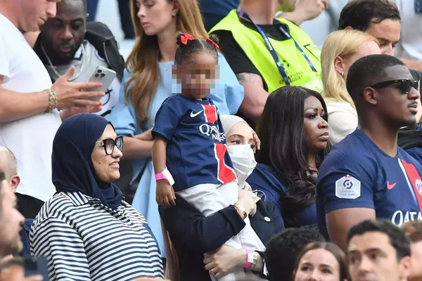 Rima Edbouche, femme d'Ousmane Dembélé, évoquée parmi des supporters du Paris Saint-Germain en tribunes, dont une femme en hijab à rayures, un enfant en maillot et une femme aux cheveux longs