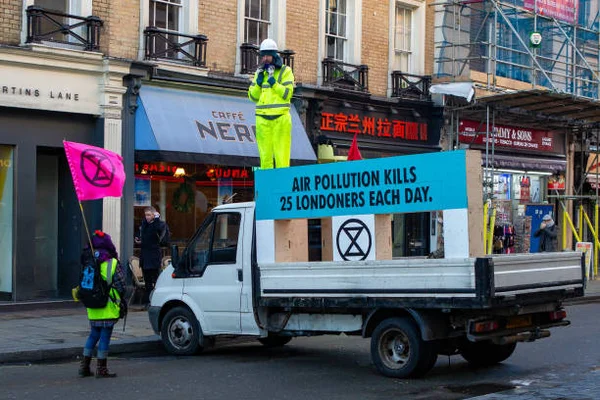 Protestation Extinction Rebellion à Londres : manifestant en gilet haute visibilité sur un camion blanc avec mégaphone, banderole bleue sur la pollution de l'air, drapeau rose XR devant un Caffè Nero