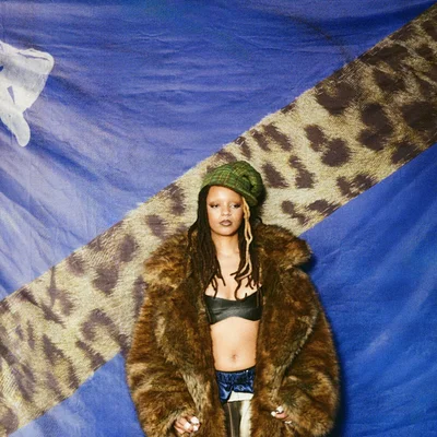Camille Yembe aux cheveux tressés, coiffée d'un bonnet vert en maille, portant un manteau en fourrure, un haut bandeau noir et un pantalon en satin bleu, devant un fond bleu à bande léopard