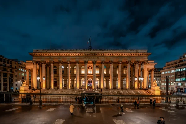 Le Palais Brongniart illuminé la nuit, avec sa colonnade néoclassique et son large escalier baignés de lumière chaude sous un ciel bleu profond, des lampadaires, un drapeau sur le toit et des piétons traversant la chaussée humide