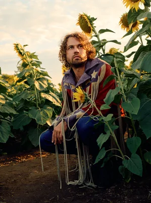 Kevin Morby, musicien barbu aux cheveux bouclés, accroupi parmi de grands tournesols à l'heure dorée, portant une chemise à franges ornée d'étoiles et regardant sur le côté
