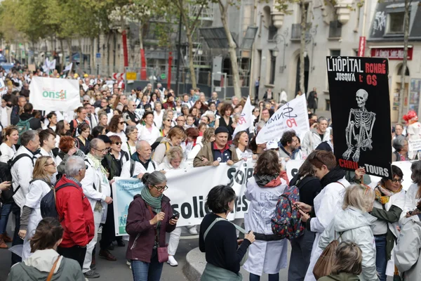 Foule dense de manifestants en blouses blanches défilant à Paris, portant des banderoles SOLIDAIRES et une grande pancarte noire L'HÔPITAL RONGÉ JUSQU'À L'OS avec un squelette, sur un boulevard bordé d'arbres d'automne