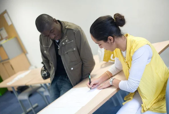 Homme en veste vert olive debout près d'une table en bois clair où une femme assise en chignon avec gilet jaune écrit sur une feuille lors d'un accompagnement pour jeunes diplômés